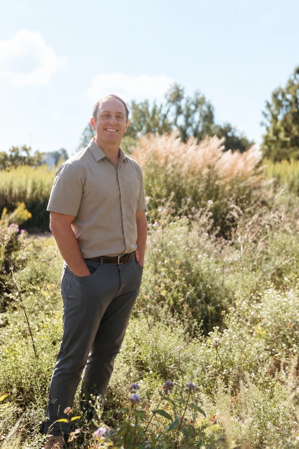 Dr. Daniel Tagge standing in a sunlit meadow of tall grass and wildflowers, smiling.
