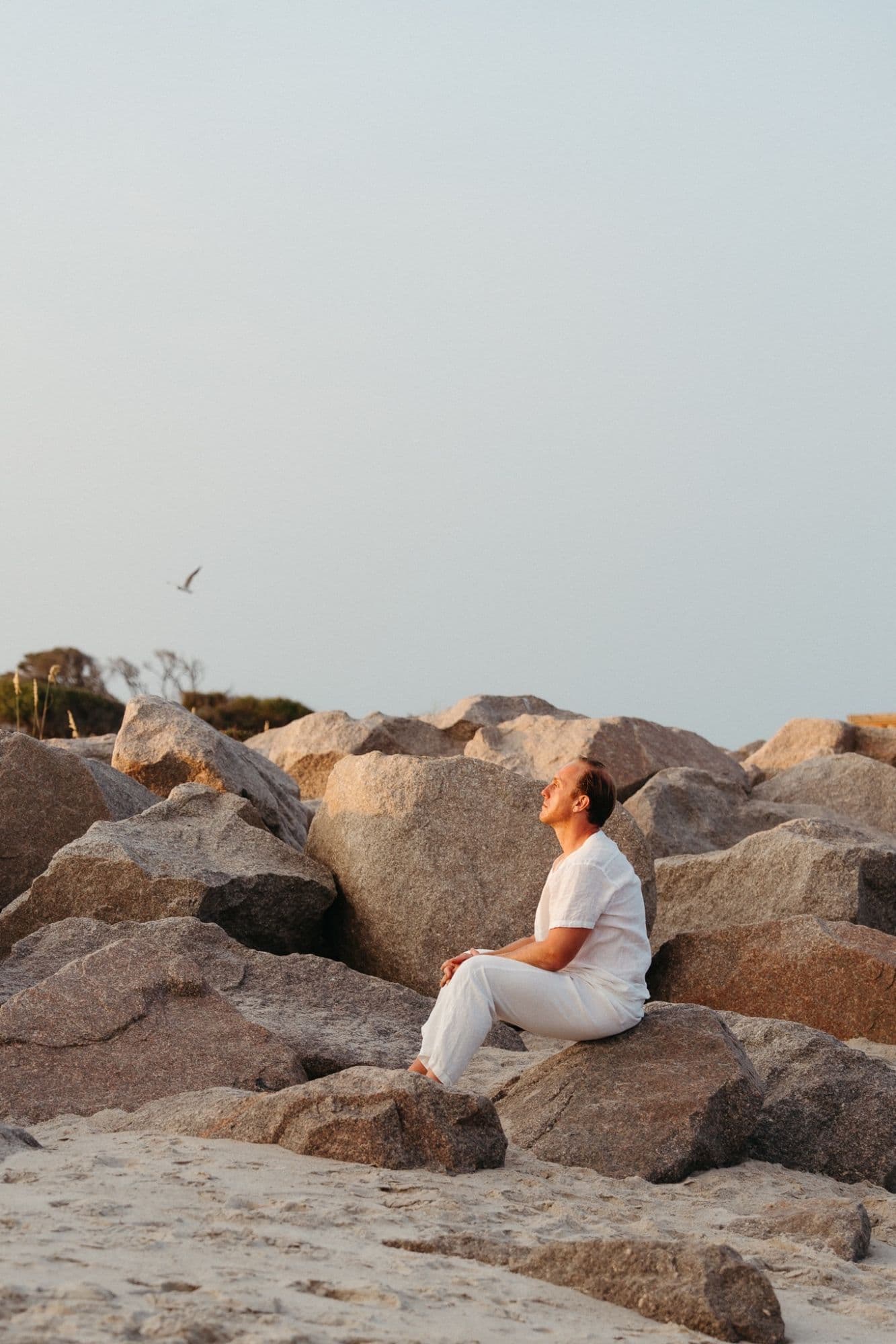 Dr. Daniel Tagge seated alone on the rocks of a jetty at the shore, looking up at the sky