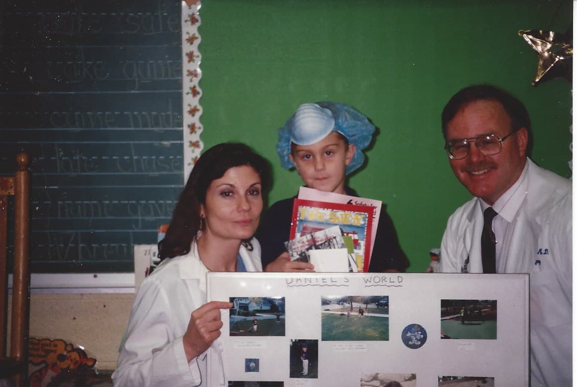 Daniel as a young boy in a surgical cap and mask in a classroom, between two adults in white coats holding a poster titled Daniel's World.