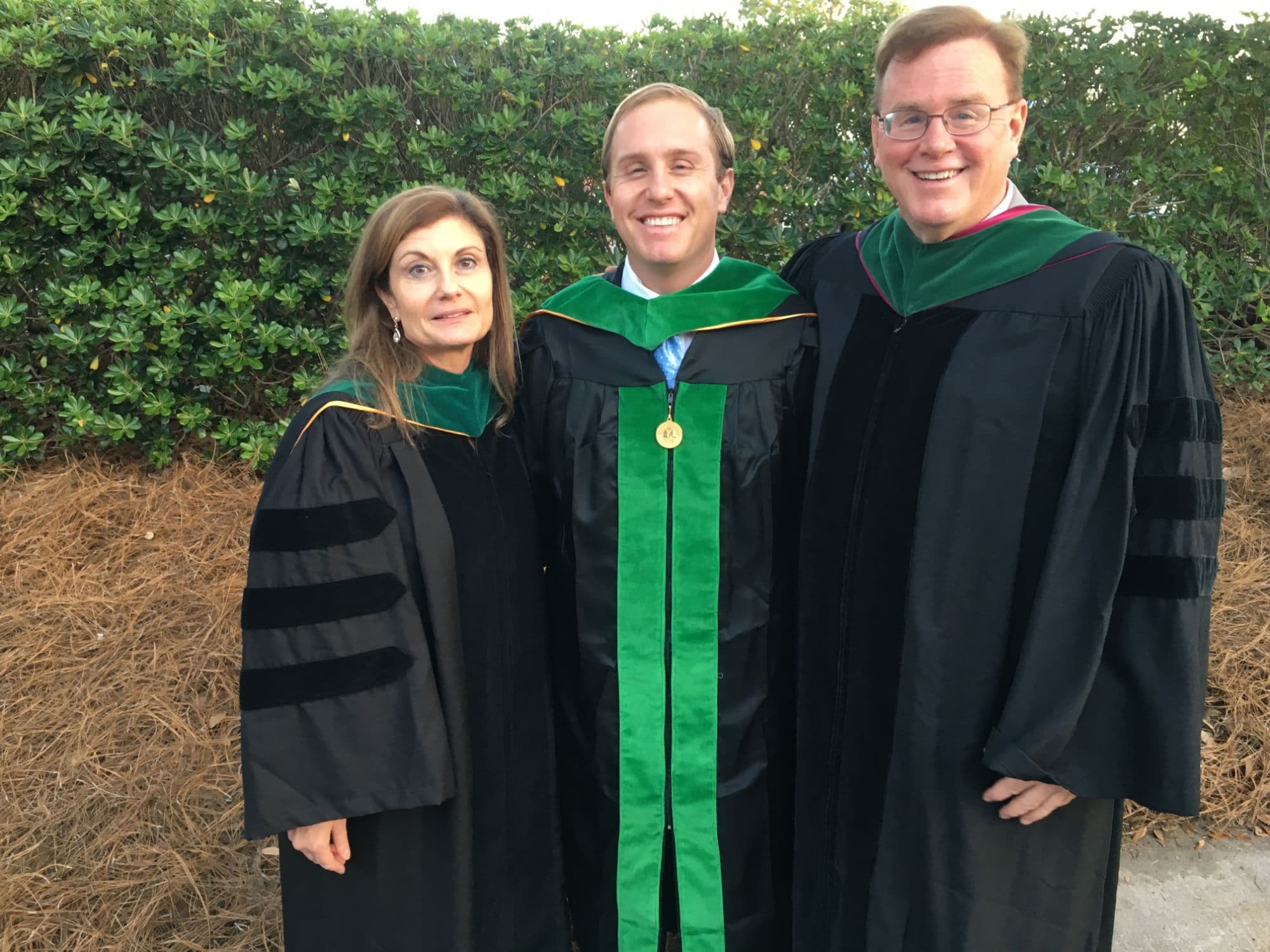 Daniel Tagge in a medical school graduation gown and green hood, standing between two adults in doctoral robes.