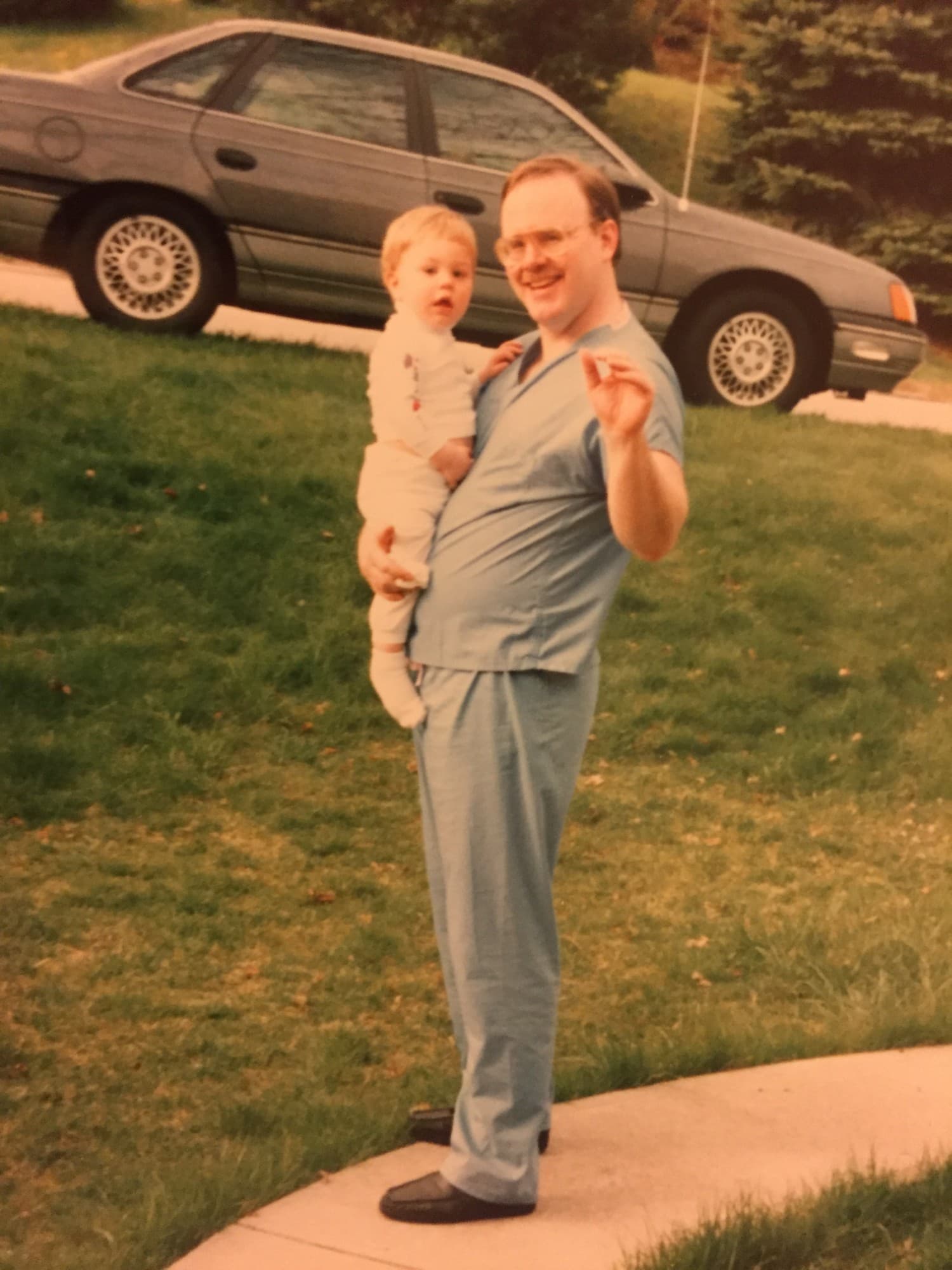 A man in blue surgical scrubs holding a toddler on his hip on a front lawn, a sedan parked behind them.